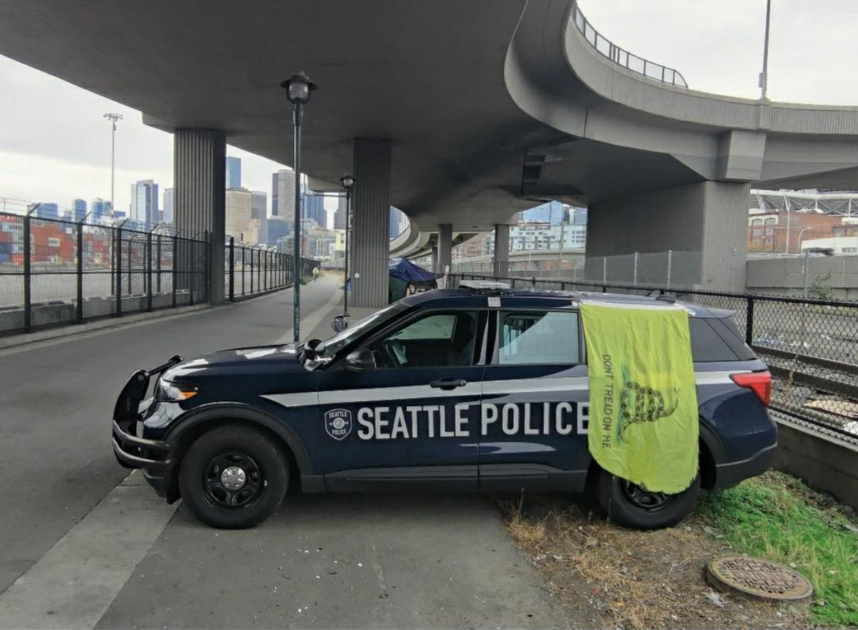 Picture of a blue Seattle Police car with a yellow Gadsden flag (a flag with a snake with the words "Don't Tread On Me" under it) hanging off of it